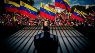 Imagem mostra líder de costas durante manifestação na Venezuela, com sombra que lembra prisão e bandeiras venezuelanas ao fundo, destacando Maduro.